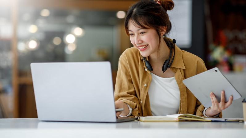 Asian woman smiling while looking at her laptop, headset around her neck.